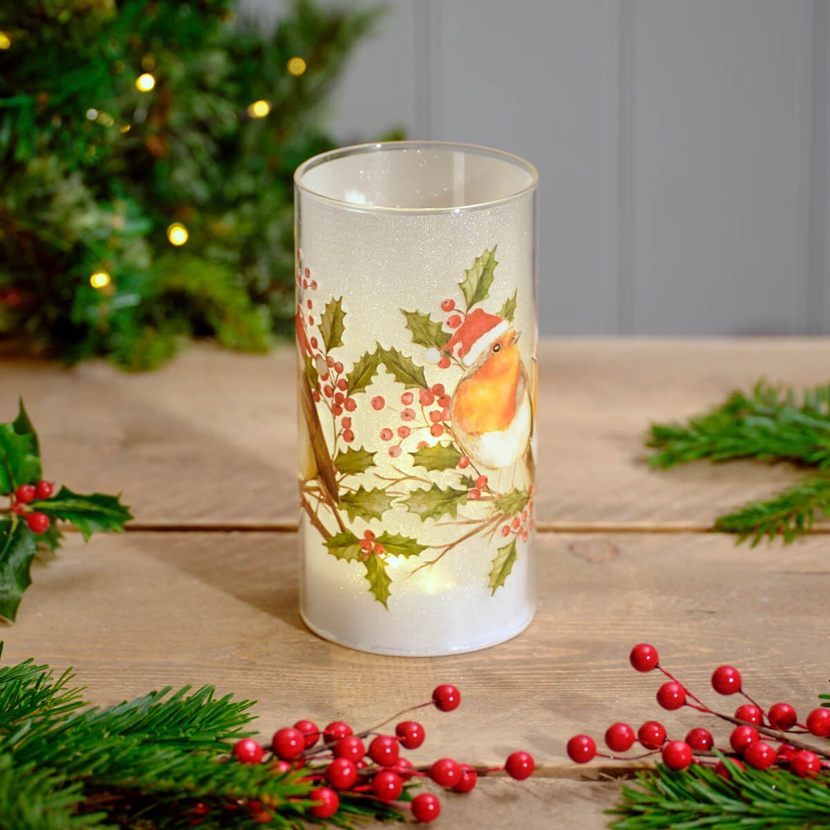 Frosted LED cylinder light with a robin and holly print glowing on a wooden table among pine and red berries