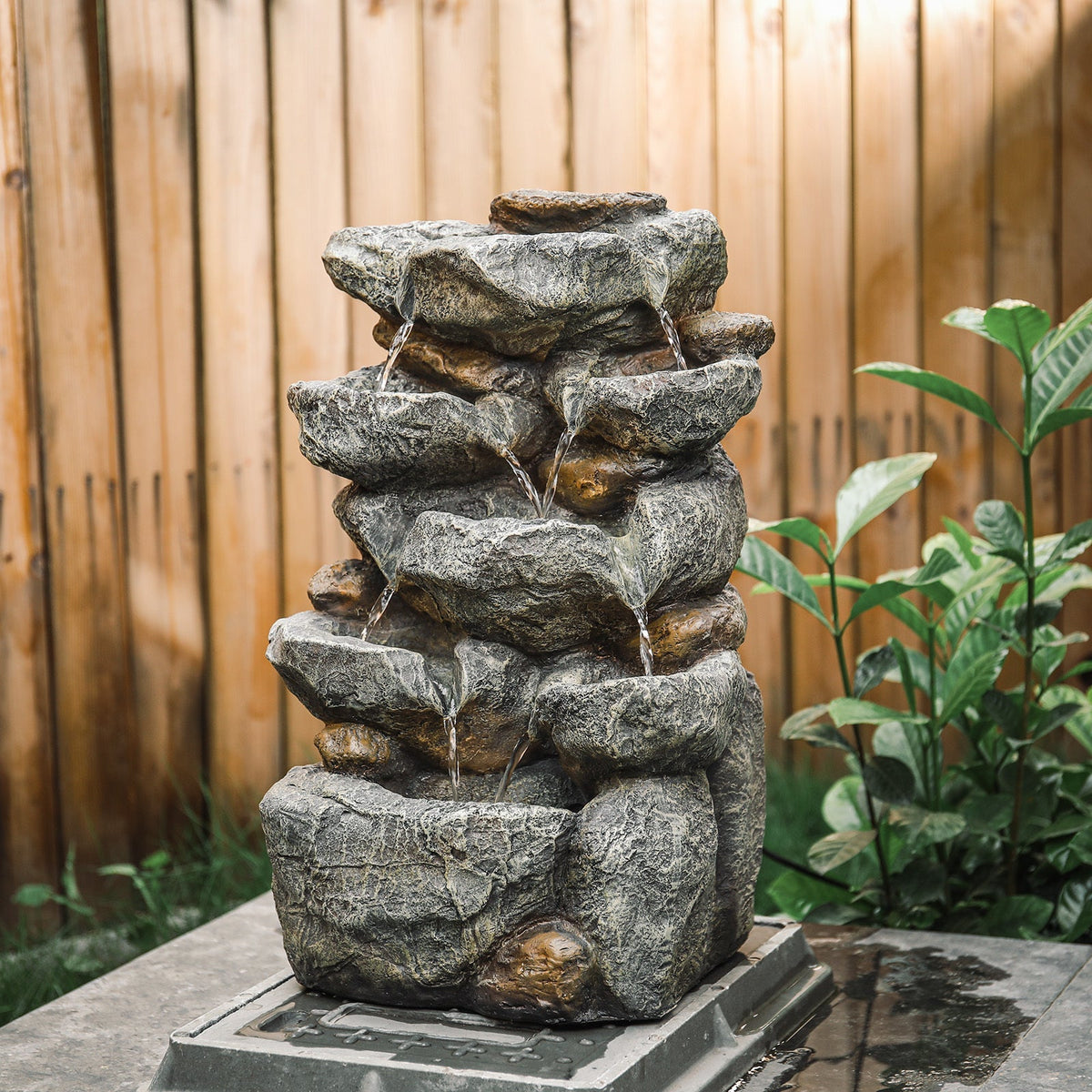 Rock cascade fountain with water running over its ledges in front of a wooden fence