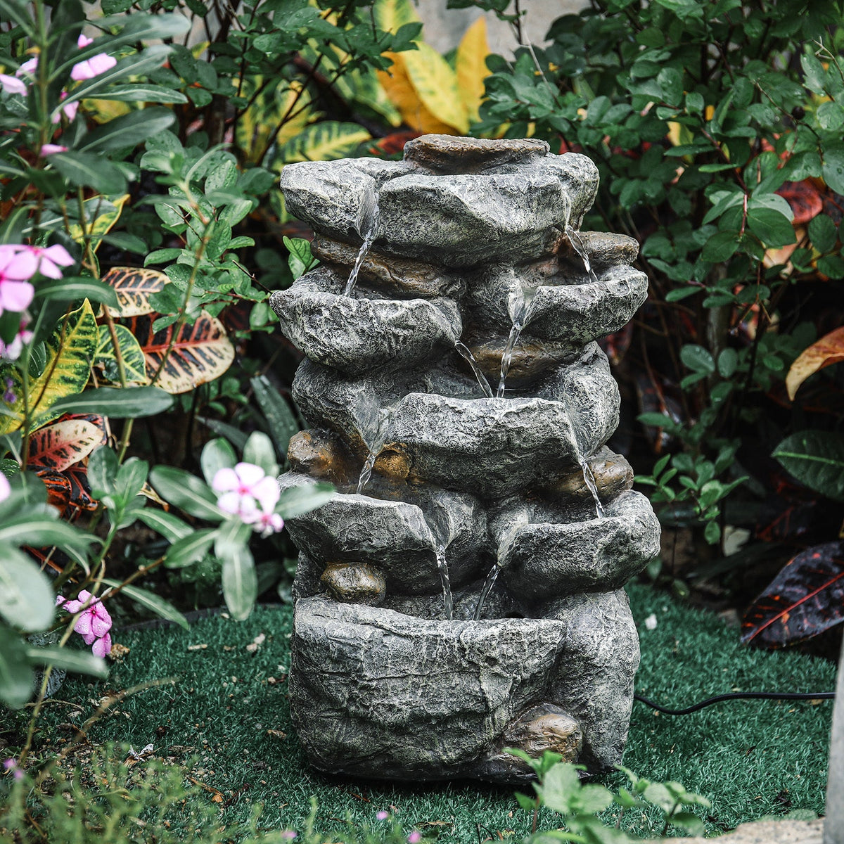 Stacked-rock water fountain among ferns and pink flowers in a garden bed