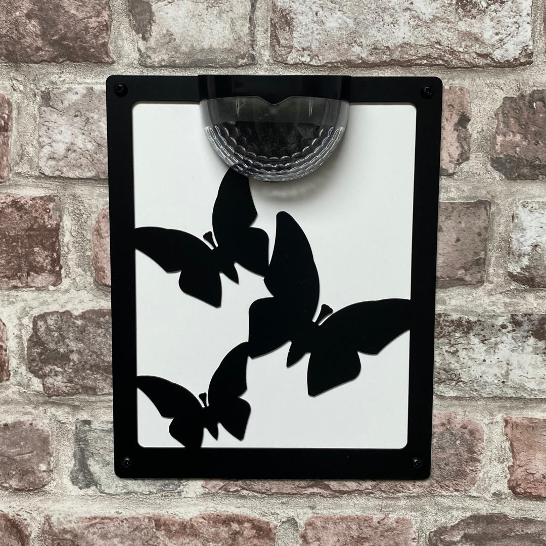 Three-butterfly solar wall plaque with white backing panel on a brick wall