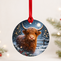 Round glass tree decoration with a shaggy Highland Cow in falling snow by a fence, on a red ribbon