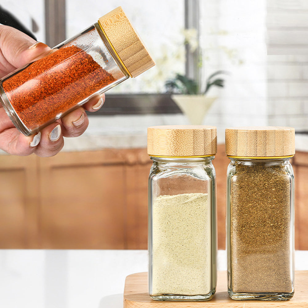 A hand shaking paprika from a glass spice jar, two filled jars with bamboo lids on a board