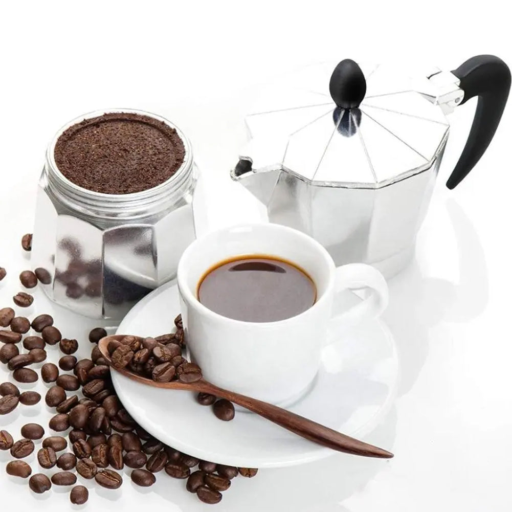 Aluminium moka pot beside an espresso cup, ground coffee and coffee beans on white
