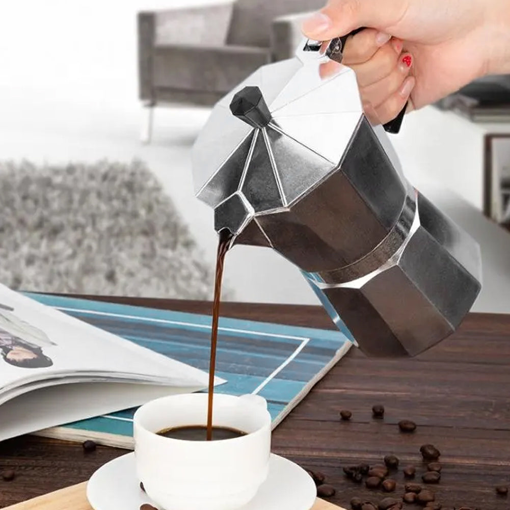 Hand pouring coffee from the eight-sided moka pot into a white cup beside coffee beans