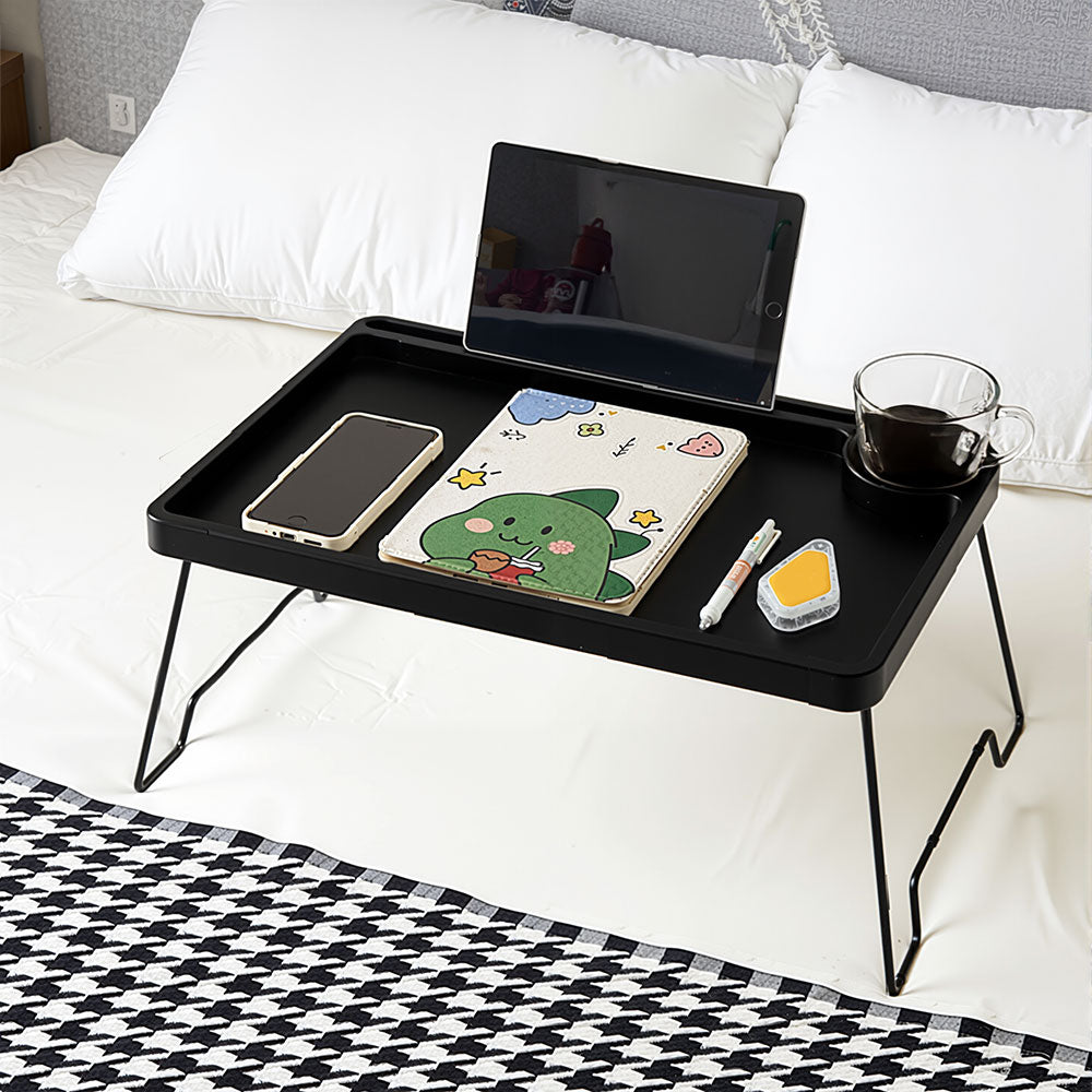 Black folding bed table on a bed with a tablet in the slot, a notebook and a drink