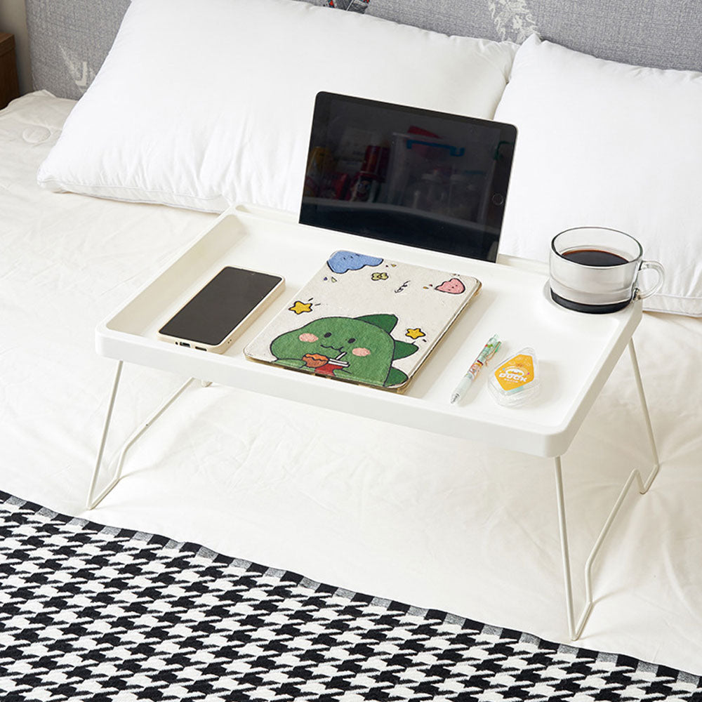 White folding bed table on a bed with a tablet in the slot and a notebook
