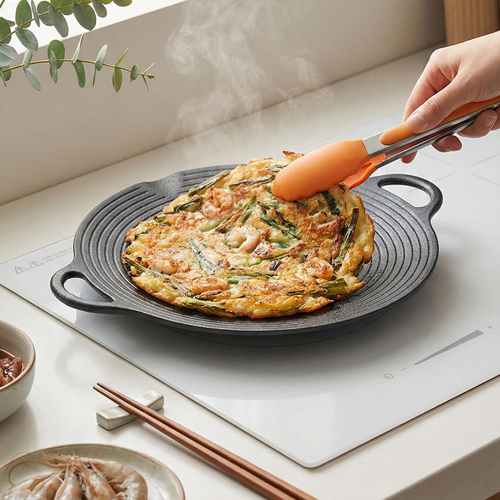 Seafood pancake cooking on the round cast iron grill pan on a white hob, lifted with orange tongs