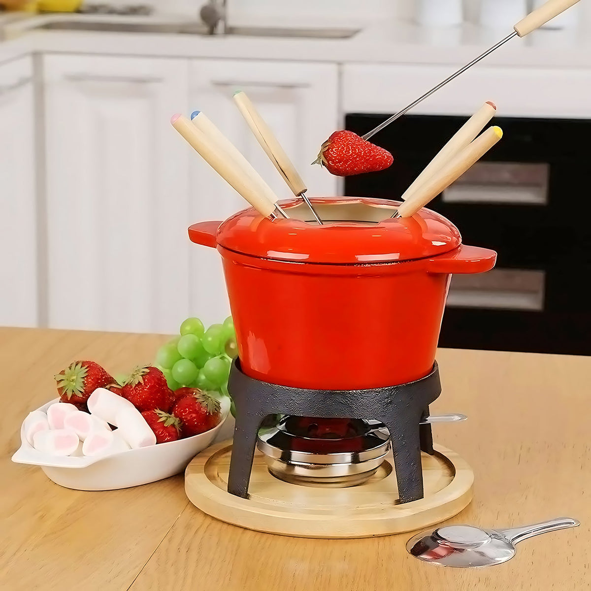 Red fondue set on its stand and wooden base with a strawberry being dipped, fruit and marshmallows beside it