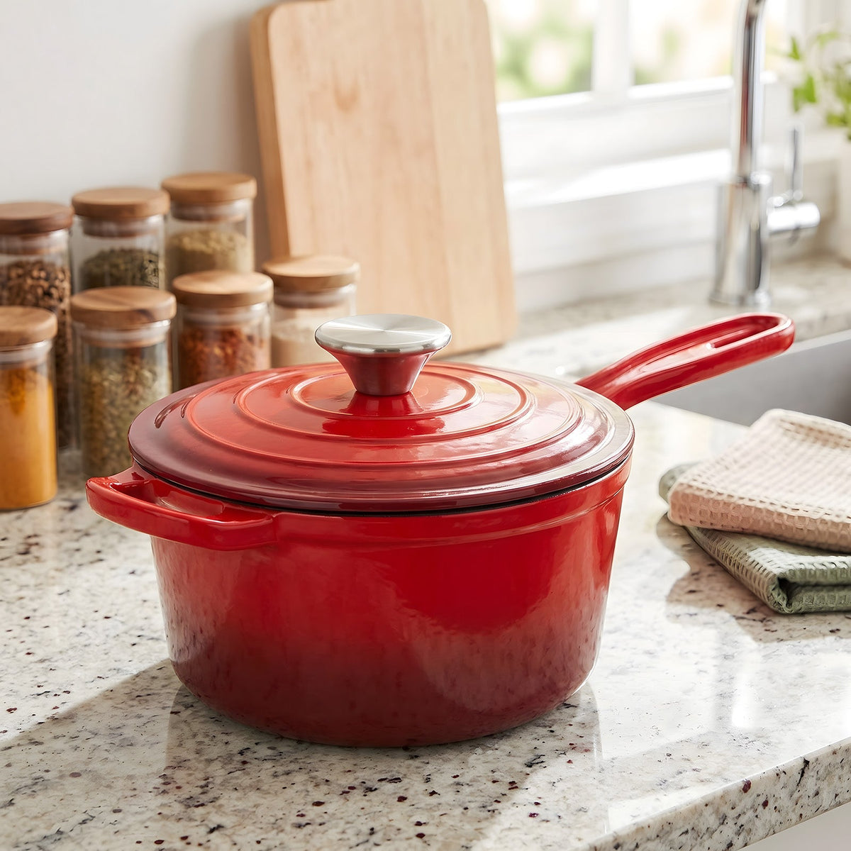 Red enamelled cast iron saucepan with its lid on, on a granite worktop by spice jars