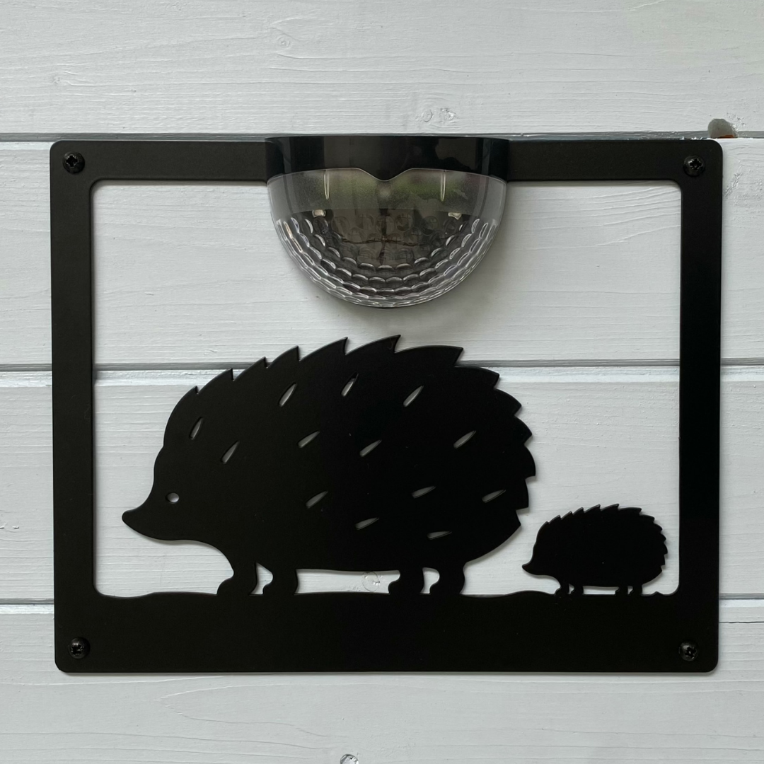 Open black hedgehog and hoglet cut-out plaque with domed solar lamp on a white painted shed wall