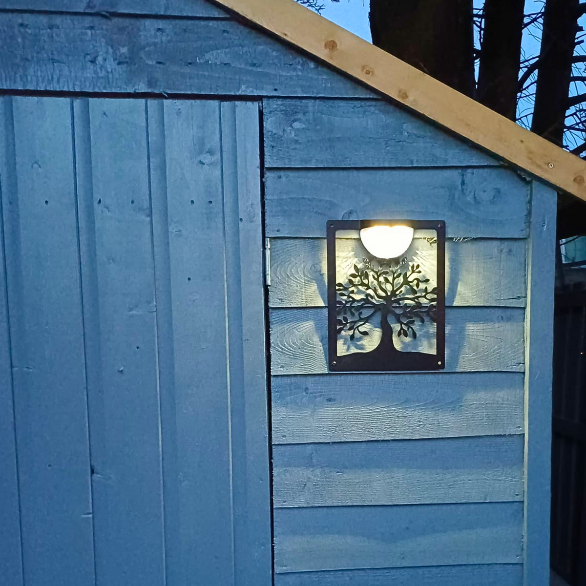 Tree of life solar plaque glowing on a blue garden shed at dusk