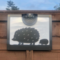 Black hedgehog and hoglet solar wall plaque with a white backing panel, fixed to a wooden fence