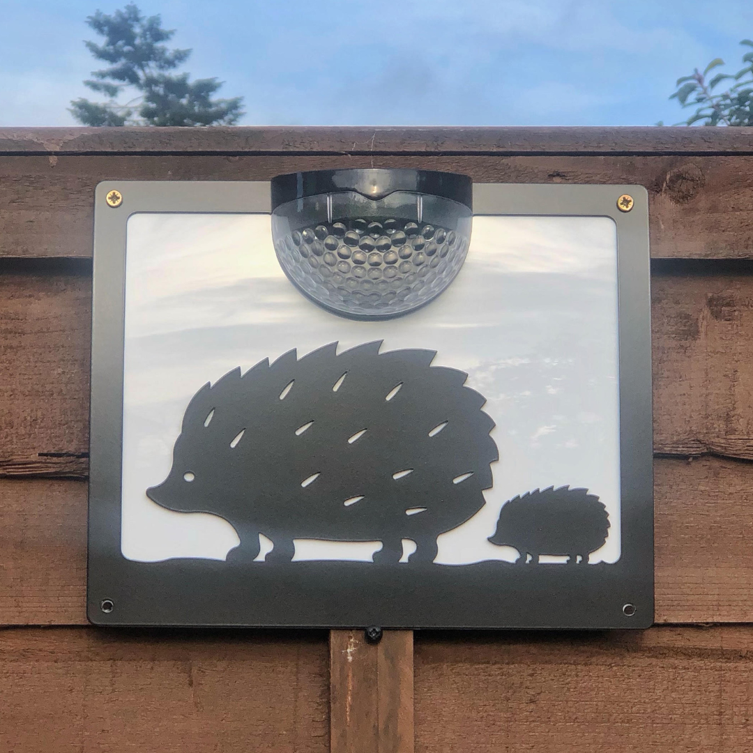 Black hedgehog and hoglet solar wall plaque with a white backing panel, fixed to a wooden fence