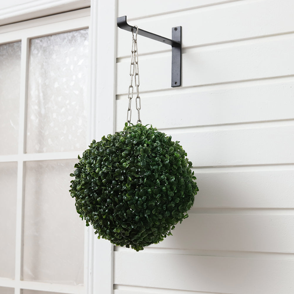 Artificial boxwood ball hanging on a chain from a black wall bracket beside a white-painted window