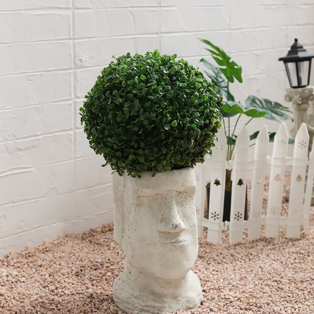 Artificial boxwood ball resting on a stone head planter on pink gravel by a white brick wall