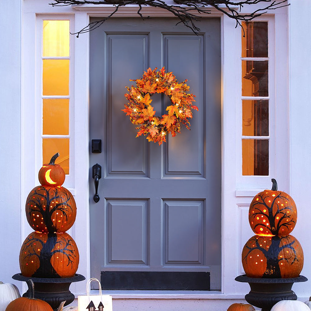 Lit maple leaf and berry wreath on a grey front door between carved pumpkins