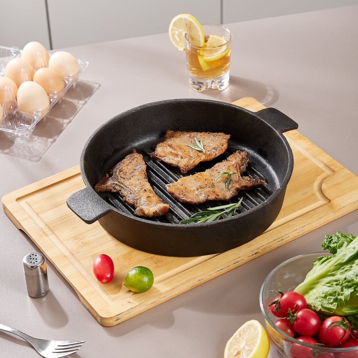 Ribbed cast iron grill pan with three pork steaks and rosemary on a bamboo chopping board