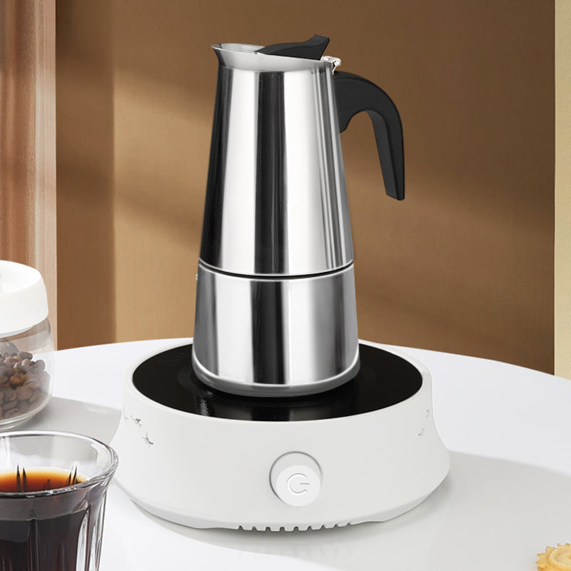 Stainless steel moka pot standing on a small white electric hotplate beside a glass of coffee