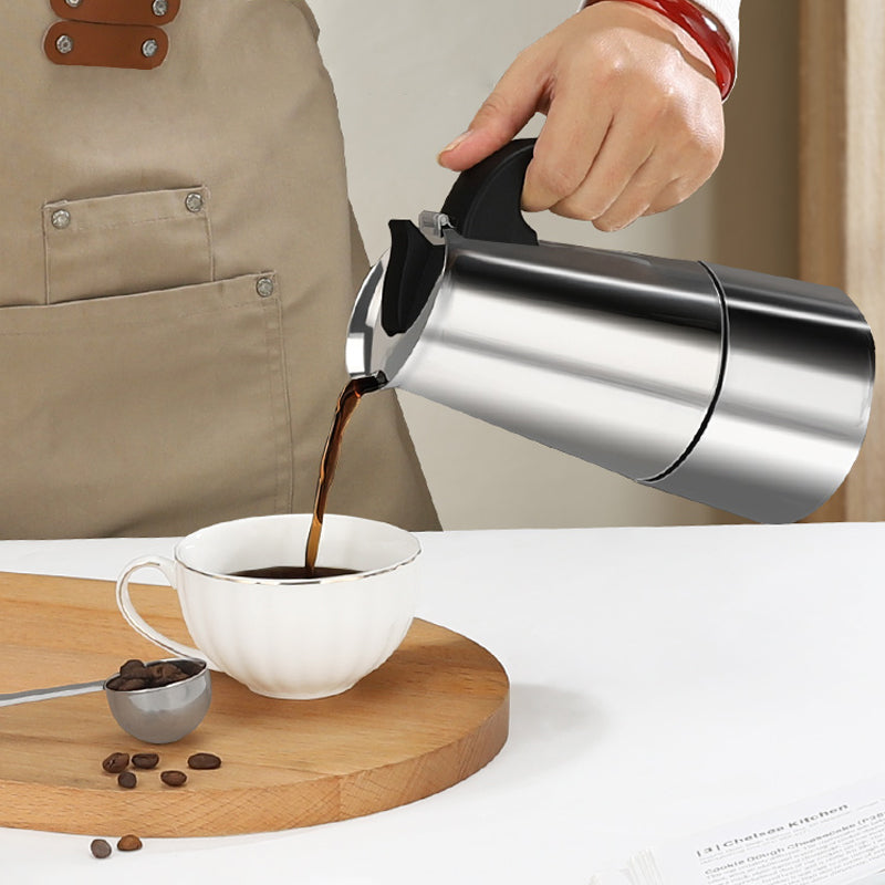 A hand pouring coffee from the stainless steel moka pot into a white cup on a wooden board