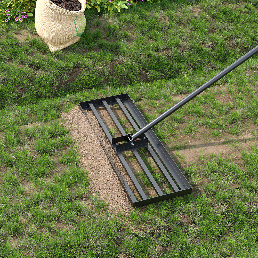 The rake spreading a band of fine topdressing over thin grass, an open hessian sack of soil and a flowering border behind it.