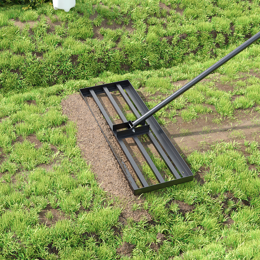 The black levelling head pulling a strip of loose soil across a patchy lawn, with the long grey handle angled away to the upper right.