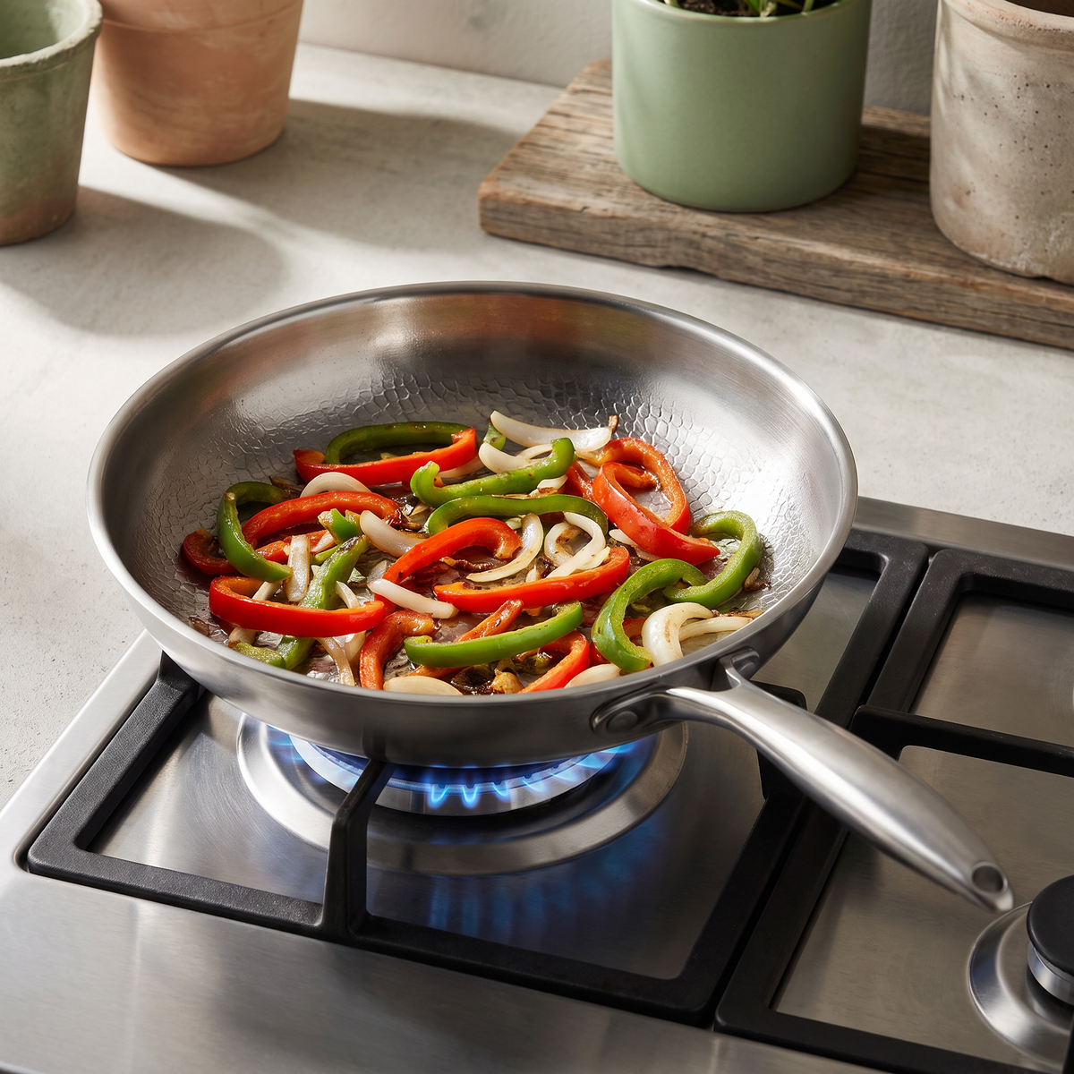 The frying pan sits over a lit blue gas flame, sliced red and green peppers and onion strips resting on its hammered interior, its long handle angled right and potted plants on the counter behind.
