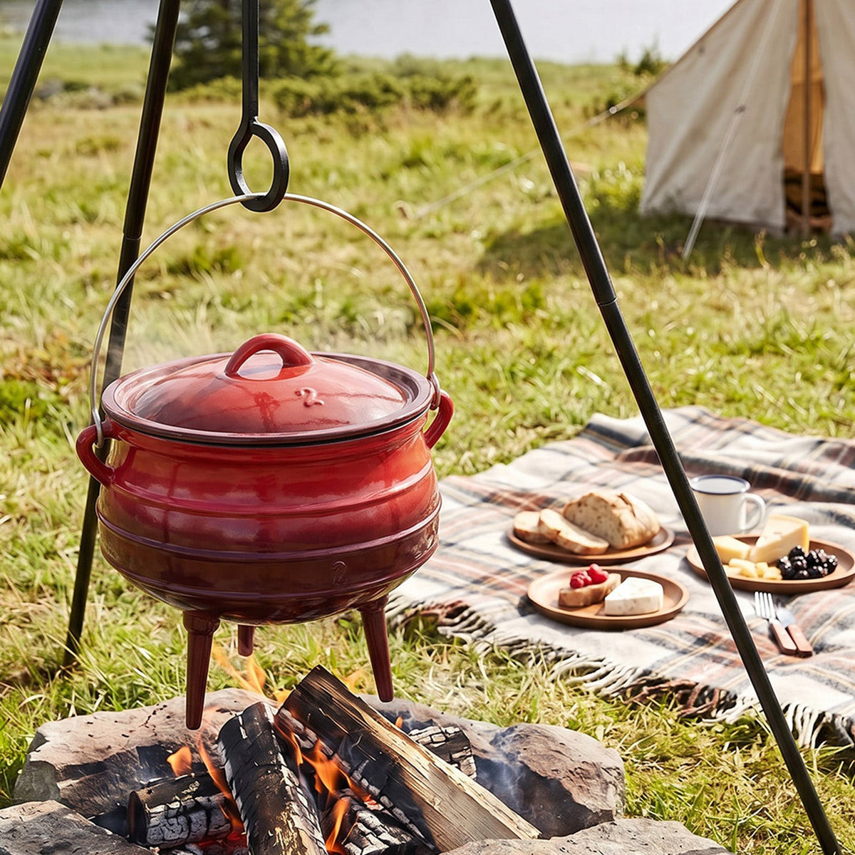 Red three-legged cast iron cauldron with its lid on, hanging from a tripod above a campfire