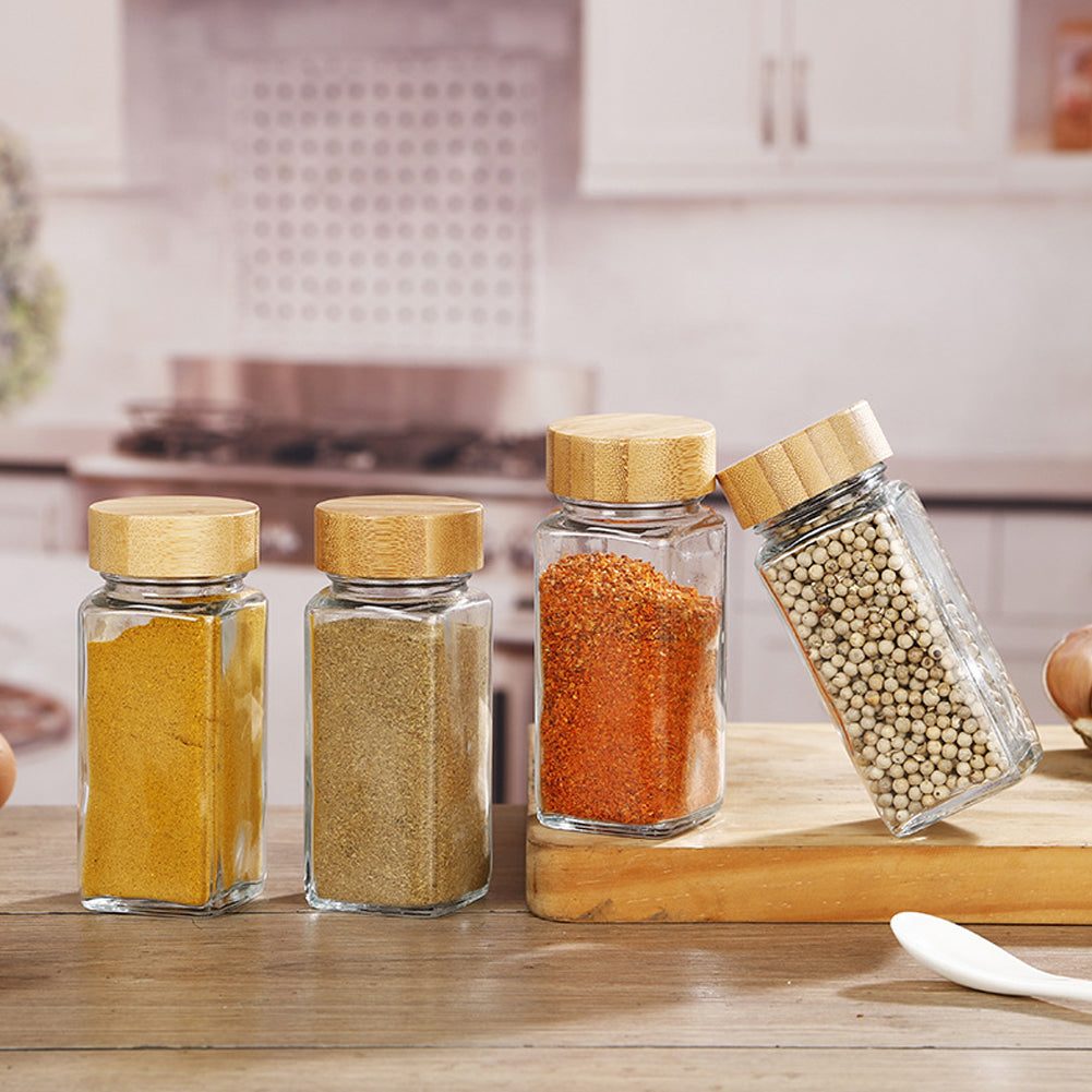 Four square glass spice jars with bamboo lids filled with turmeric, herbs, paprika and peppercorns on a kitchen worktop