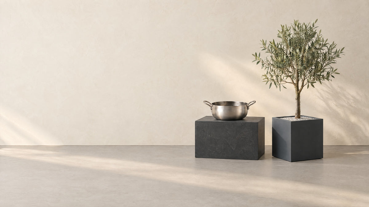 A hammered titanium stock pot on a charcoal stone block beside a square charcoal planter with a small olive tree, against a cream plaster wall