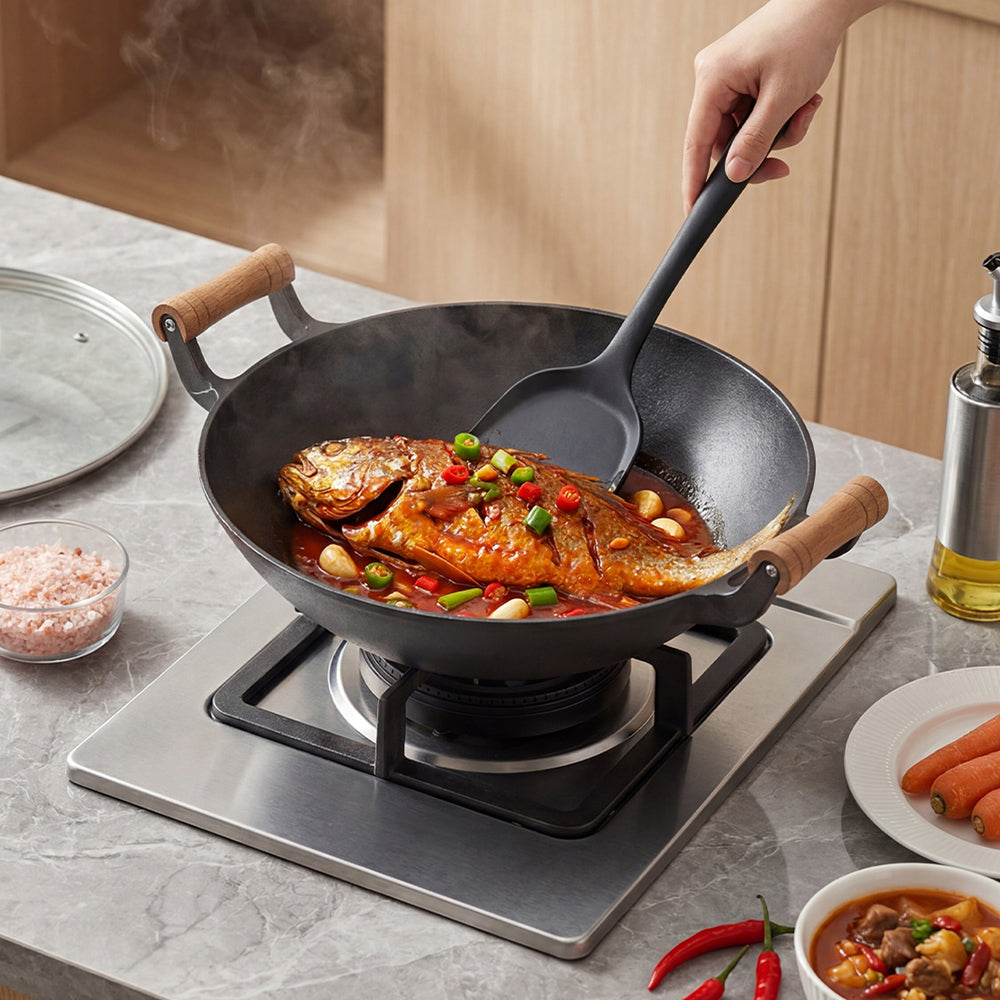 Whole fish in sauce cooking in the cast iron wok, a hand turning it with a black spatula