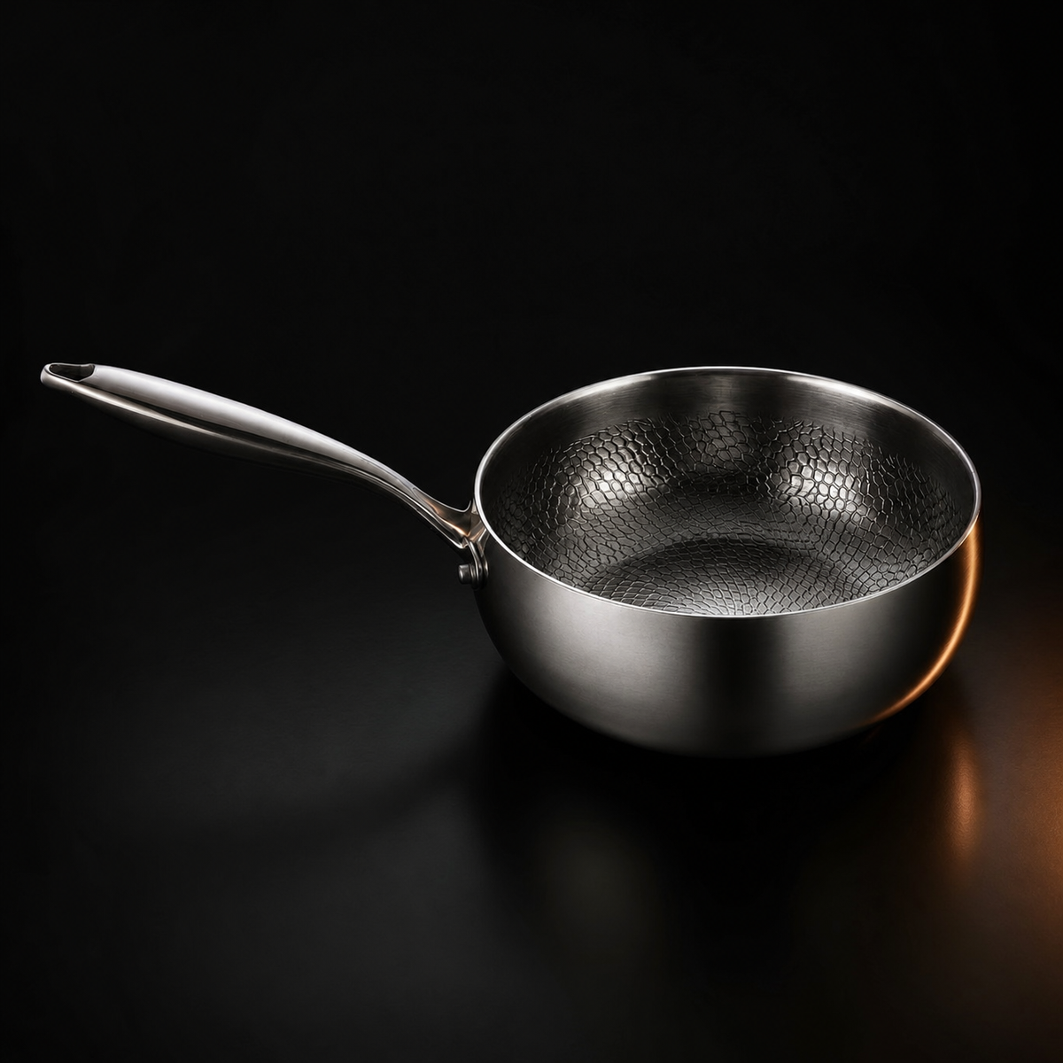 A brushed titanium saute pan with a long handle and a hammered honeycomb interior, photographed on a black background.