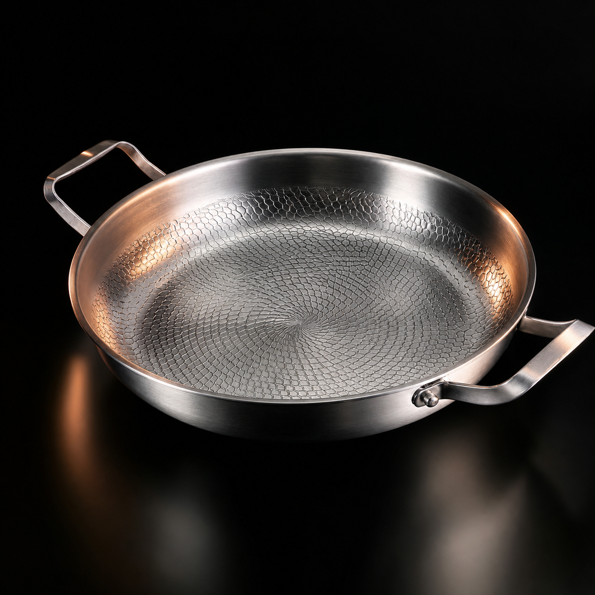 A two-handled titanium frying pan with a hammered honeycomb interior, lit against a black reflective background.