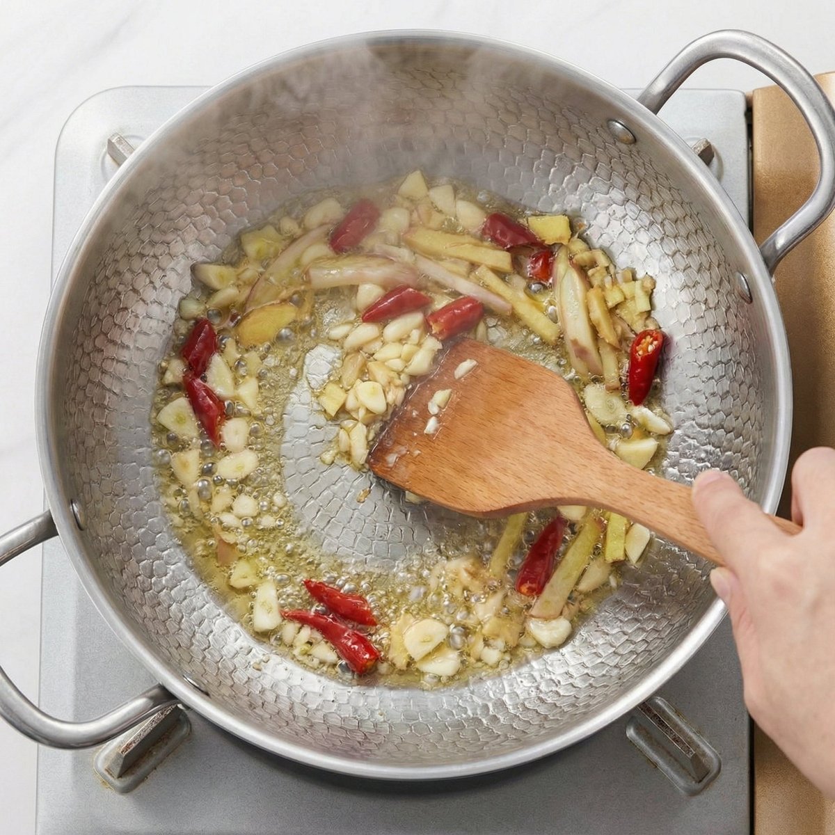 Overhead view of garlic, ginger and dried chillies frying in oil in the hammered pot, pushed around with a wooden spatula.