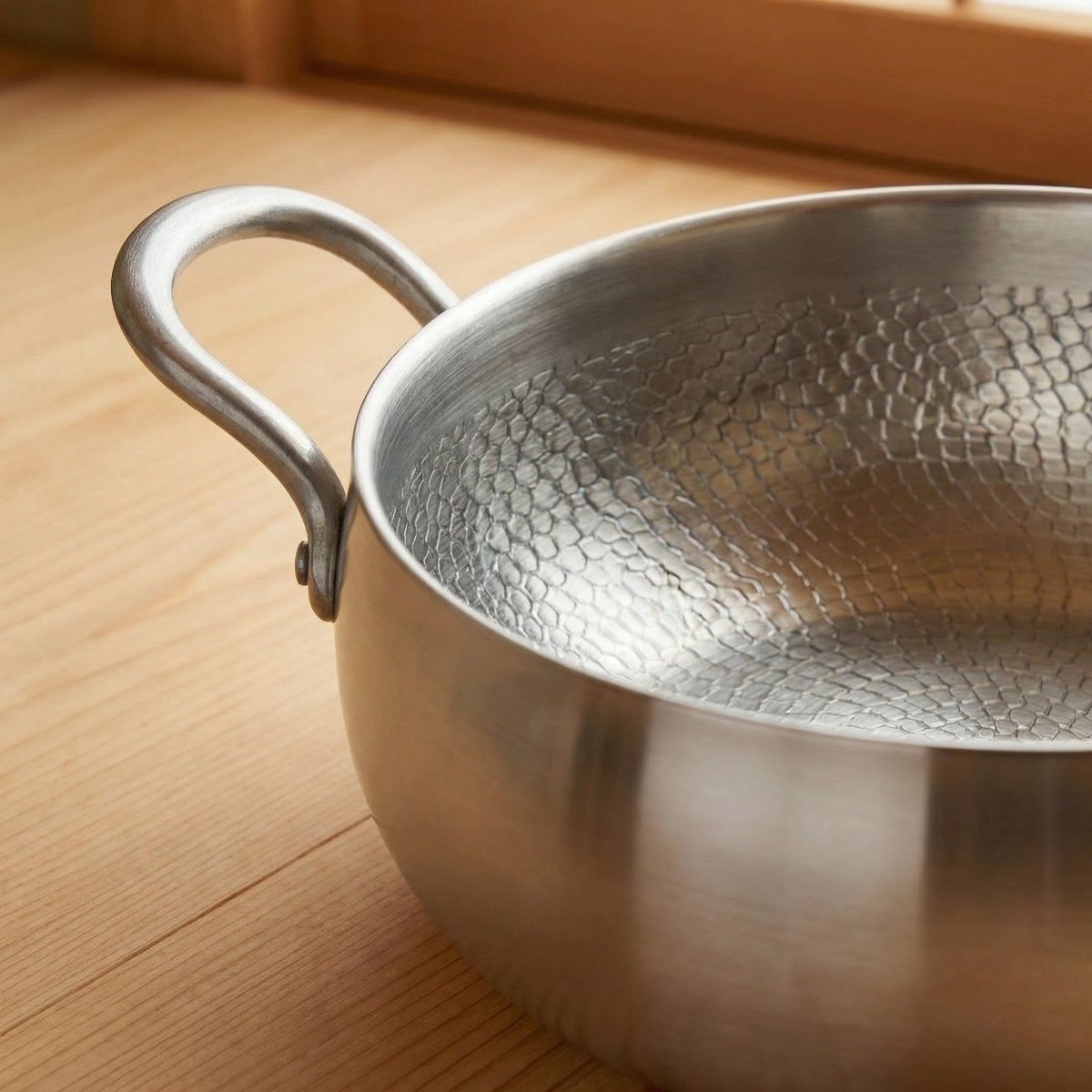 Close-up of the pot's riveted loop handle and the hammered texture inside the rim, on a pale wooden floor by a window.