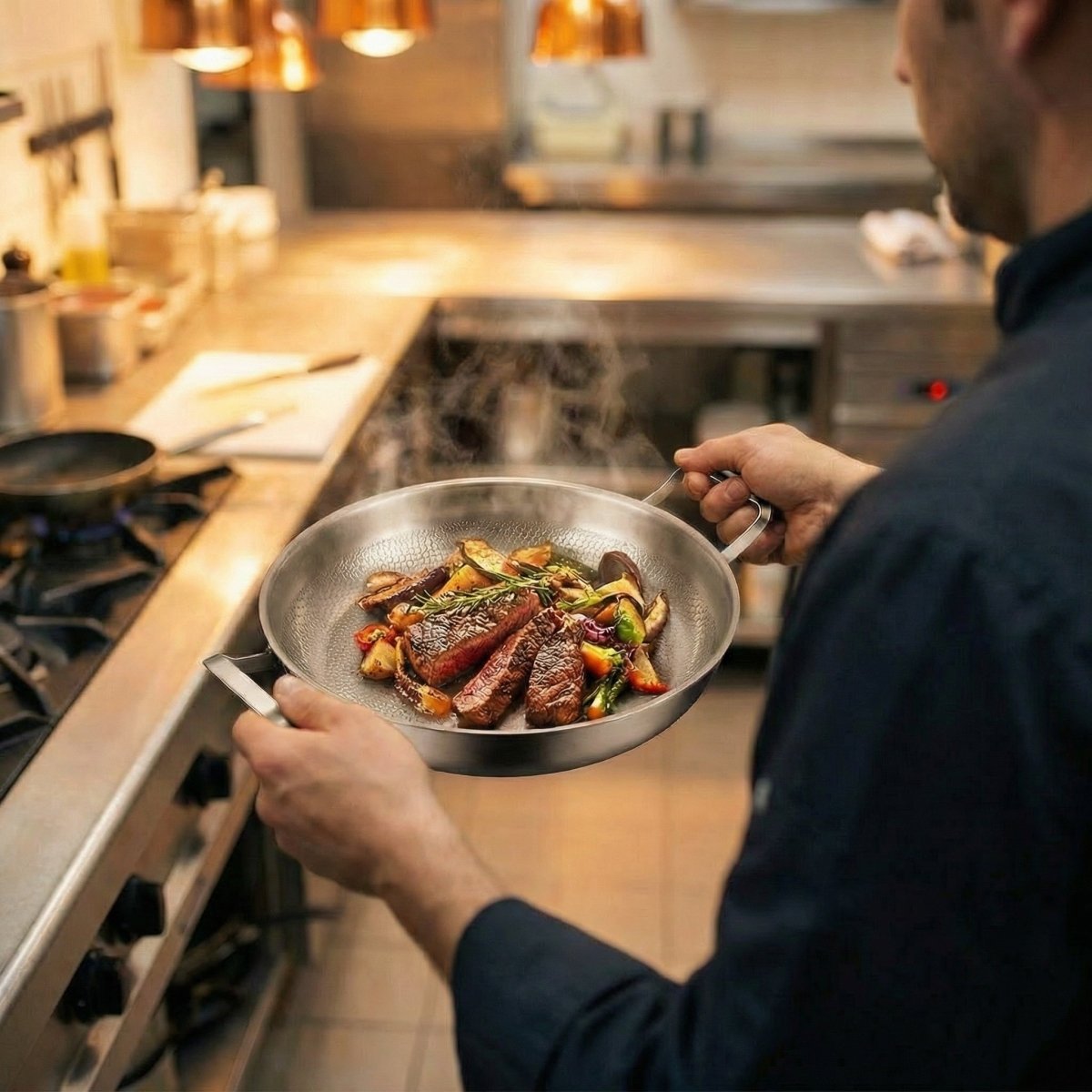 A cook in a dark jacket holds the pan by both handles, steak slices and vegetables inside, in a restaurant kitchen.
