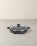 Round black cast iron braiser with two loop handles and a clear glass lid on a pale concrete floor
