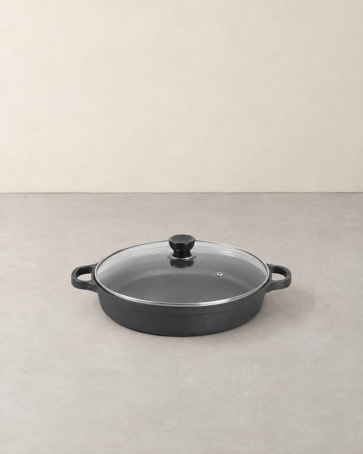 Round black cast iron braiser with two loop handles and a clear glass lid on a pale concrete floor