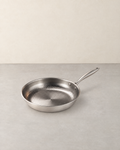 Hammered titanium frying pan with a long handle standing empty on a pale concrete floor against a cream wall