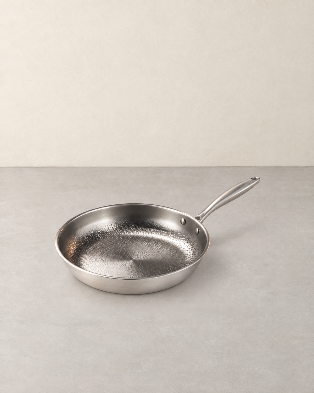 Hammered titanium frying pan with a long handle standing empty on a pale concrete floor against a cream wall