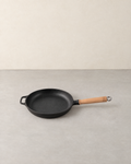 Round black cast iron skillet with a long wooden handle and a helper loop on a pale concrete floor