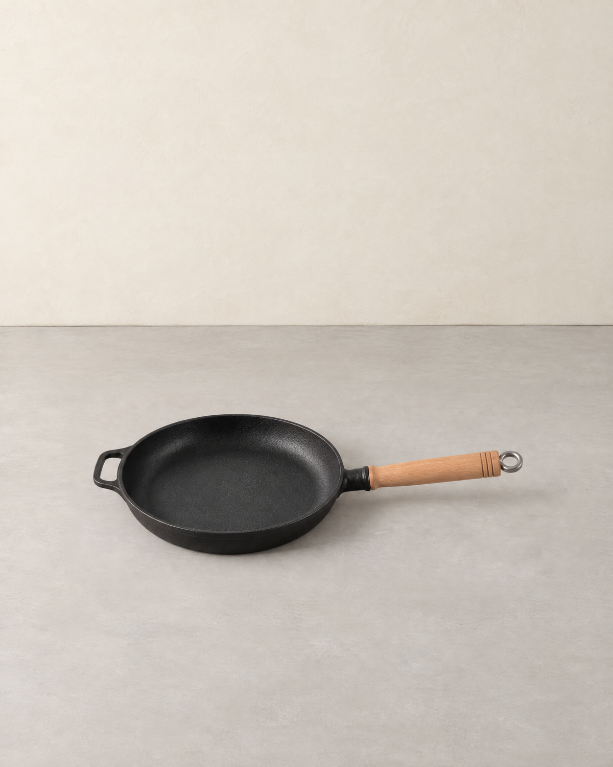 Round black cast iron skillet with a long wooden handle and a helper loop on a pale concrete floor