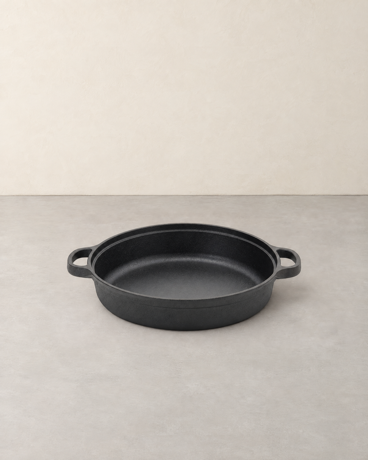 Wide shallow black cast iron skillet with two side loop handles on a pale concrete floor