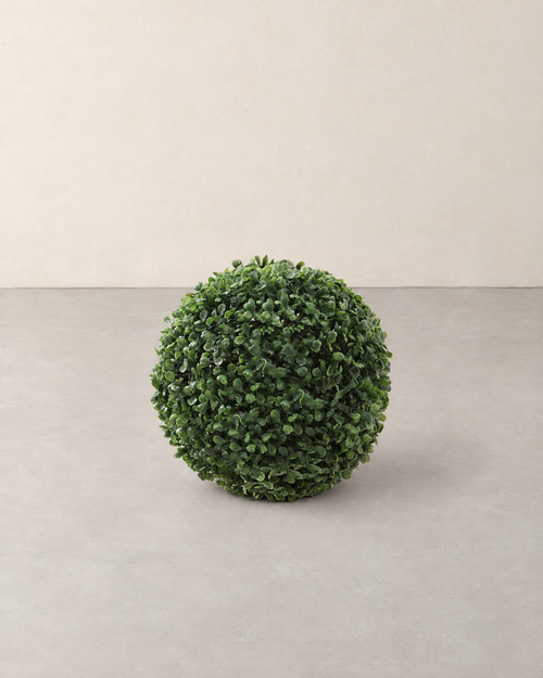 Round green artificial boxwood topiary ball standing on a pale grey floor against a cream wall
