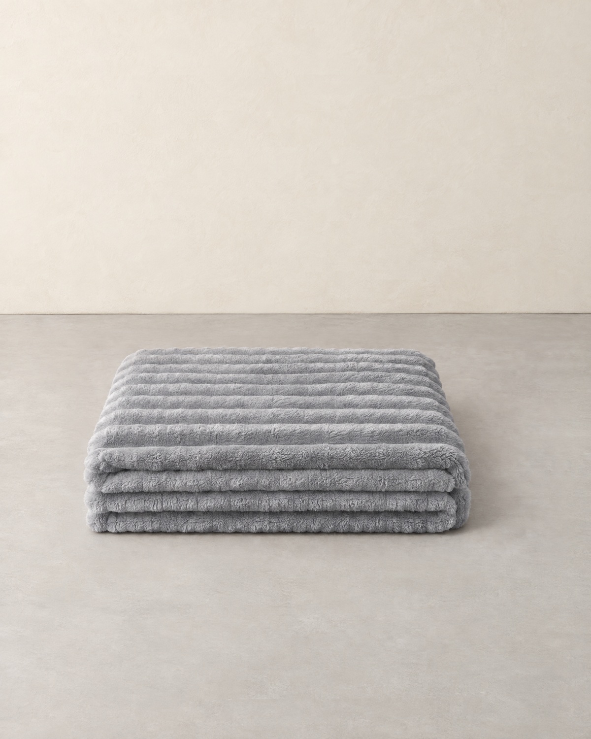 Light grey ribbed plush throw blanket folded on a pale grey floor against a cream wall