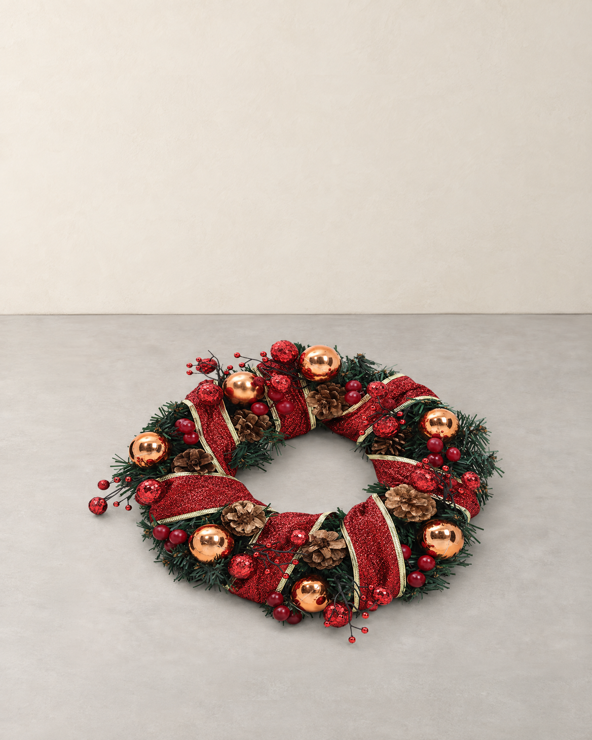 Christmas wreath with red glitter ribbon, copper baubles and pine cones lying on a pale grey floor against a cream wall
