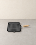 Square ribbed cast iron grill pan with a folding wire and wooden handle on a pale concrete floor