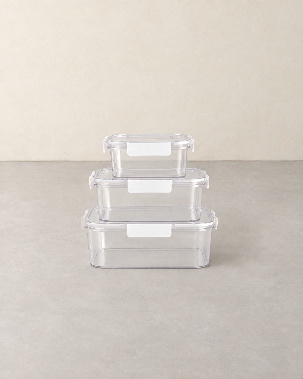 Three clear food storage boxes with white clip lids stacked largest to smallest on a pale concrete floor