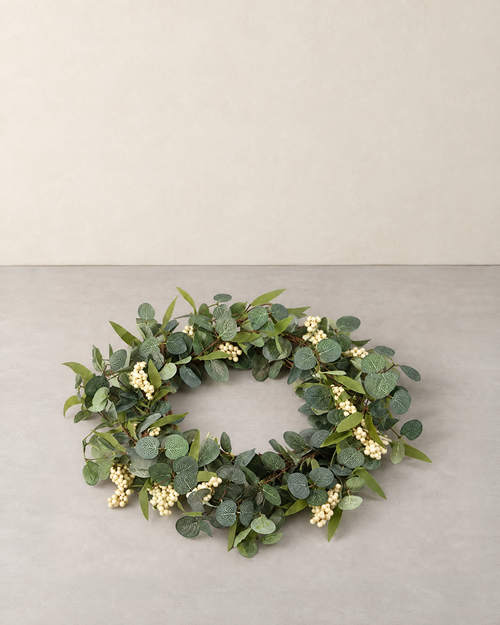 Artificial eucalyptus wreath with clusters of white berries lying flat on a pale grey floor against a cream wall