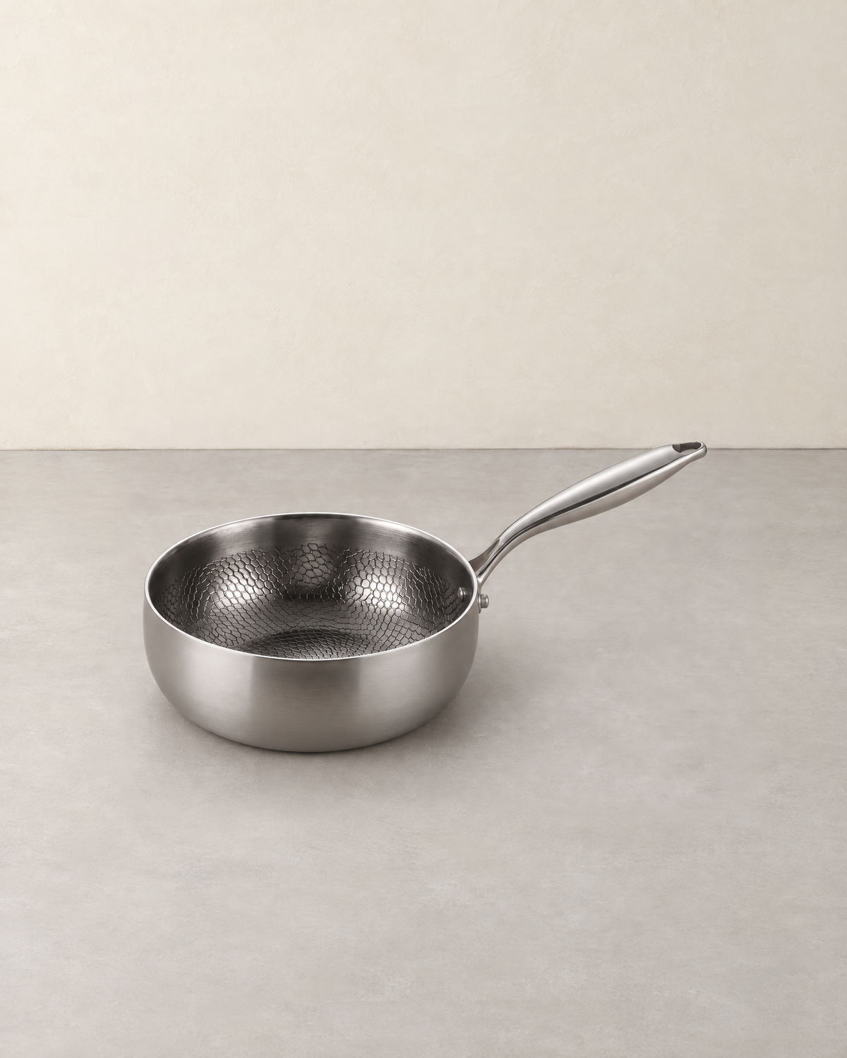 Deep hammered titanium saute pan with a long handle standing empty on a pale concrete floor against a cream wall