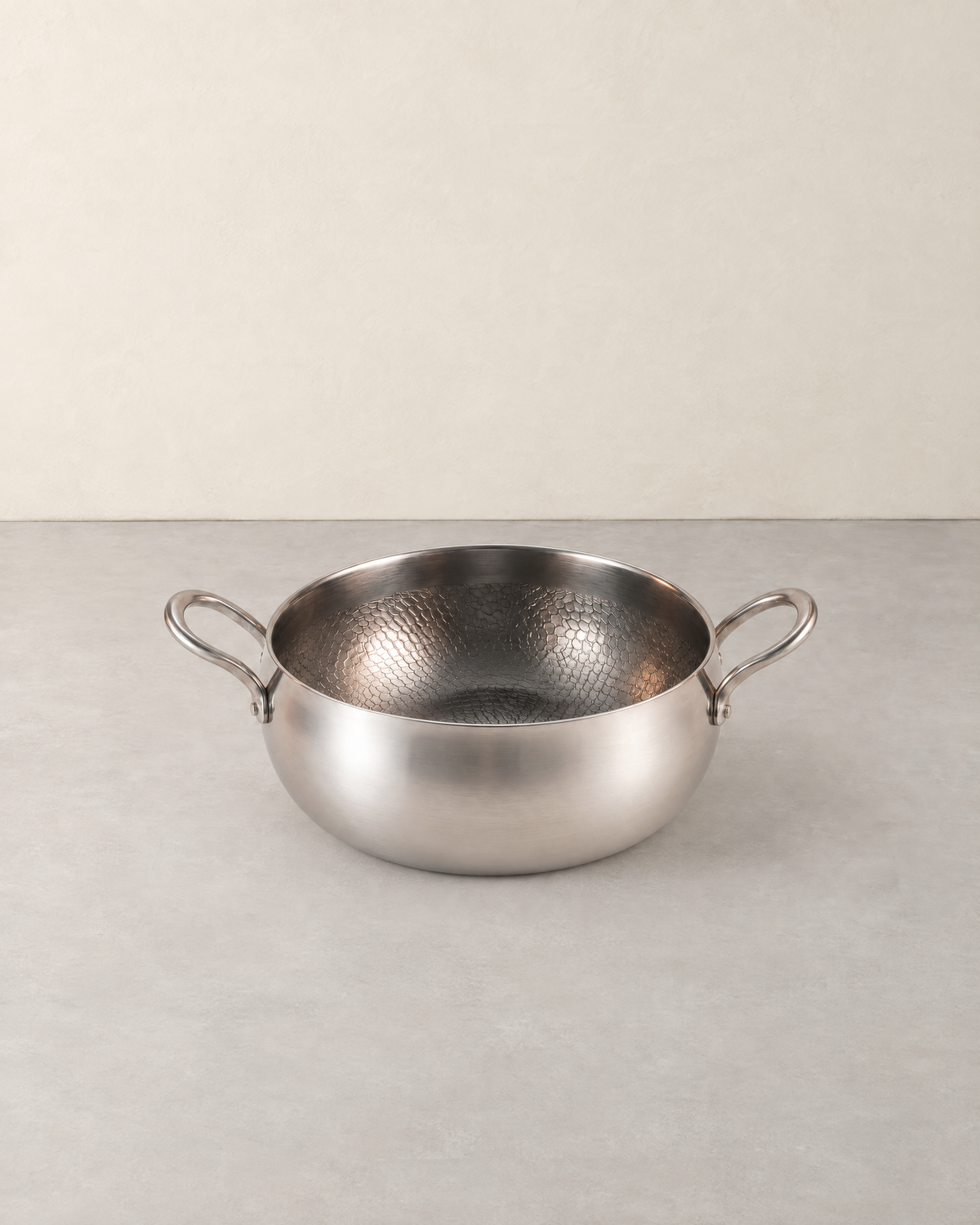 Deep hammered titanium stock pot with two loop handles standing empty on a pale concrete floor against a cream wall
